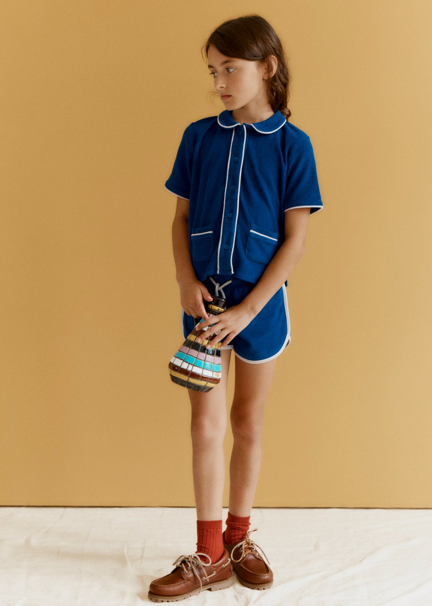 Young girl in a blue outfit holding a striped bag against a beige background