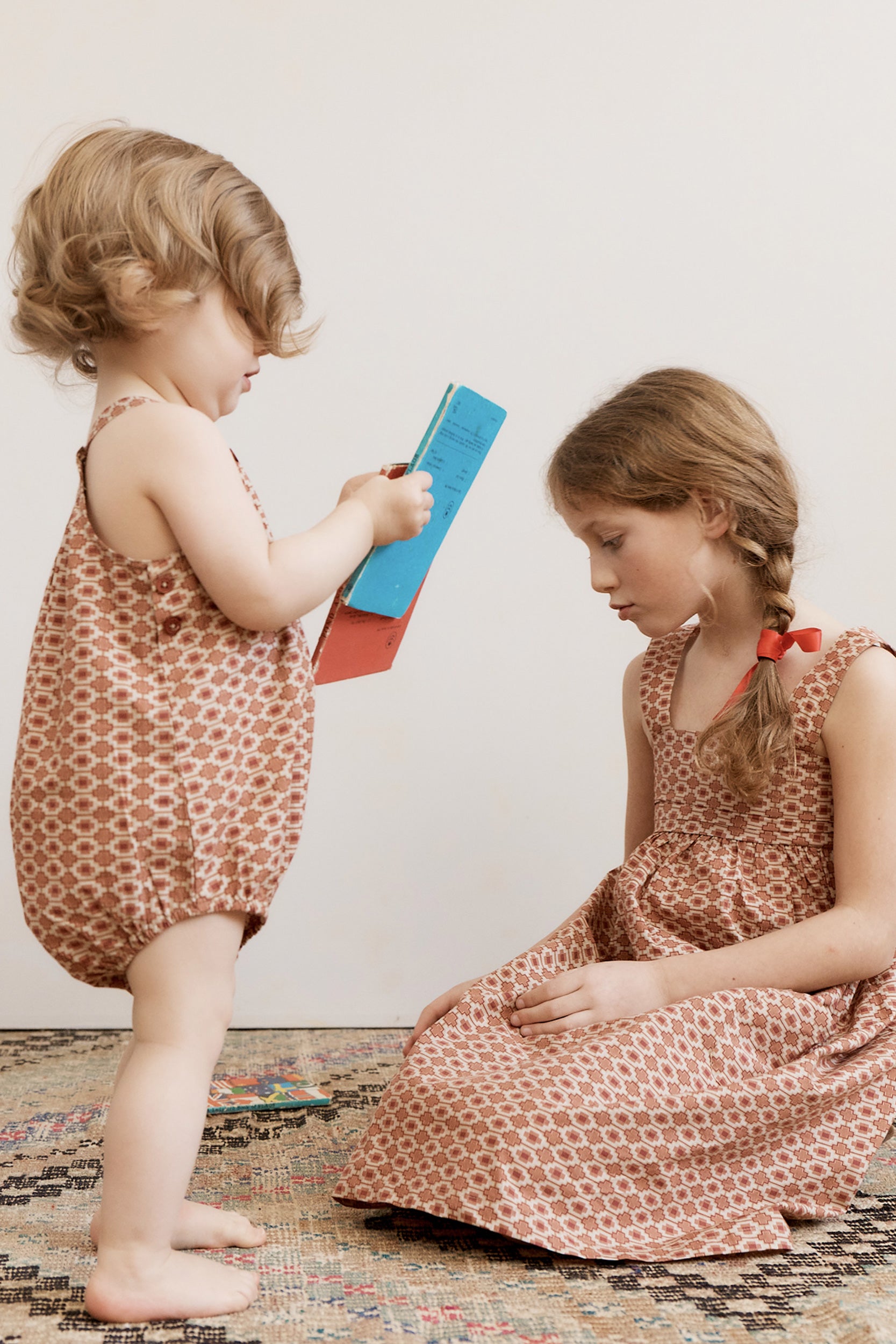 Two children in matching outfits playing with a book on a patterned rug.