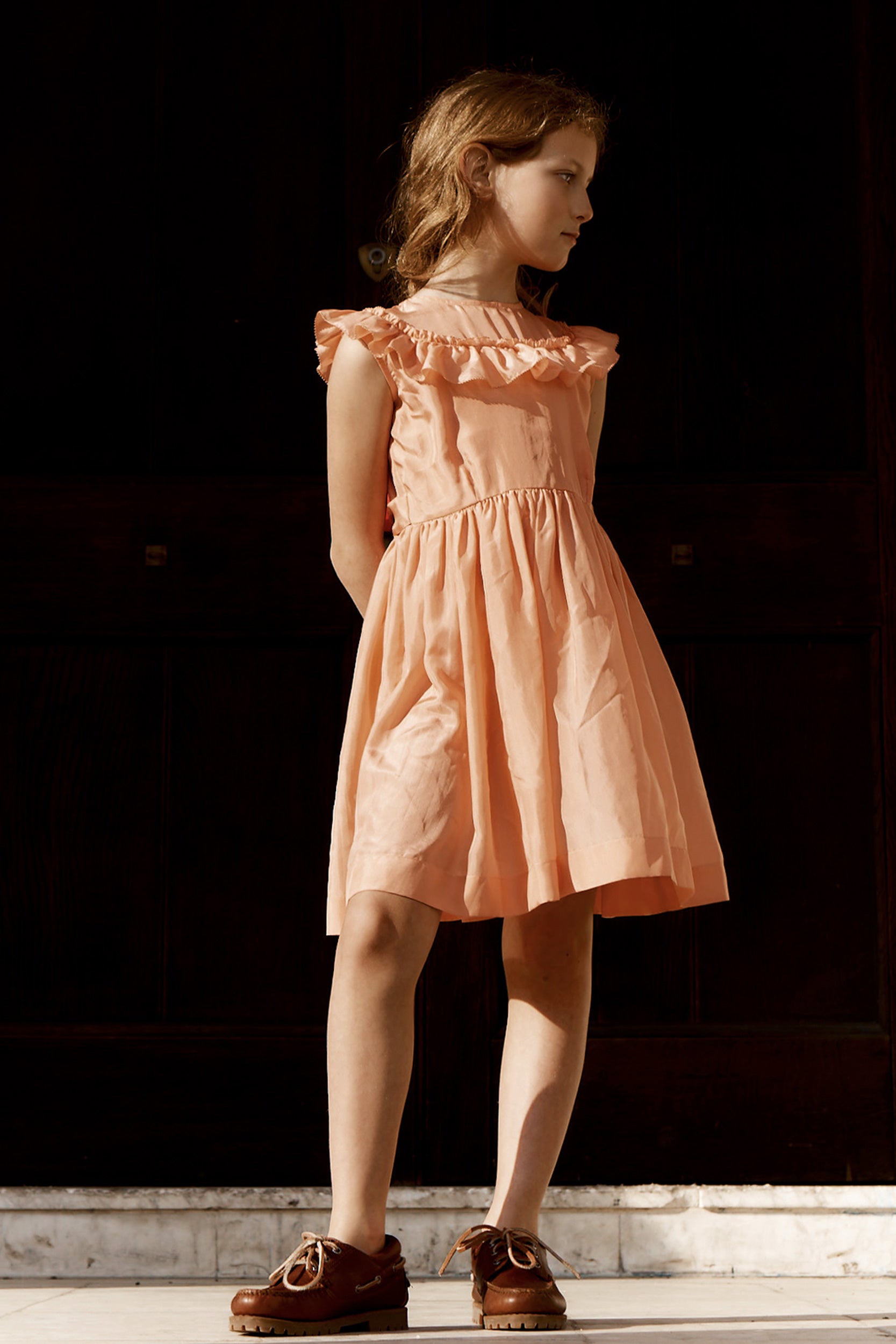 Young girl wearing a peach dress standing against a dark background